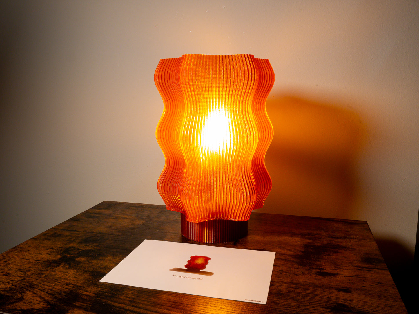 The REEF Table Lamp (Amber) & Card Gift Set emits a gentle glow on a wooden table, paired with a card featuring its image—perfect for any romantic occasion.