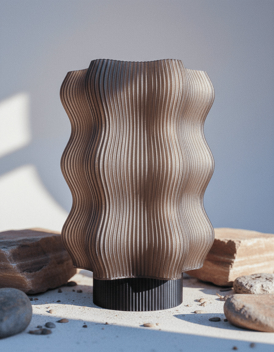 A wavy, ribbed, brown vase with a matte finish sits on a surface surrounded by rocks and stones, lit by natural light.