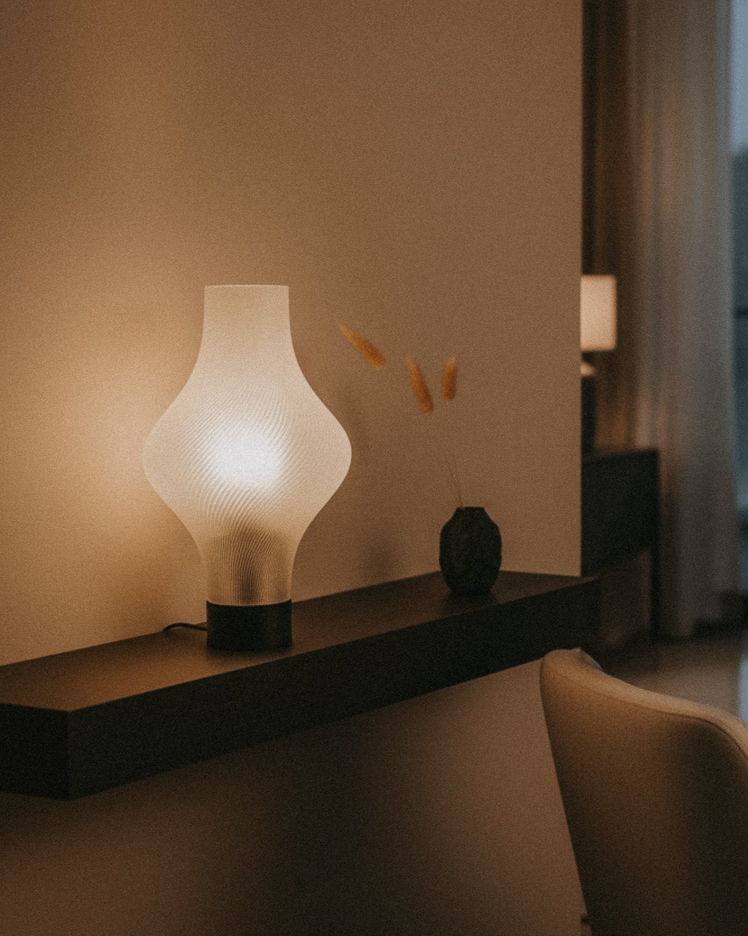 A modern white table lamp sits on a floating dark shelf next to a small vase with dried grass in a softly lit, minimalist room.