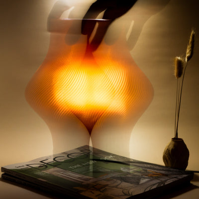 A blurred hand adjusts a modern, glowing orange lamp beside a stack of magazines and a small ceramic vase with dried grasses.