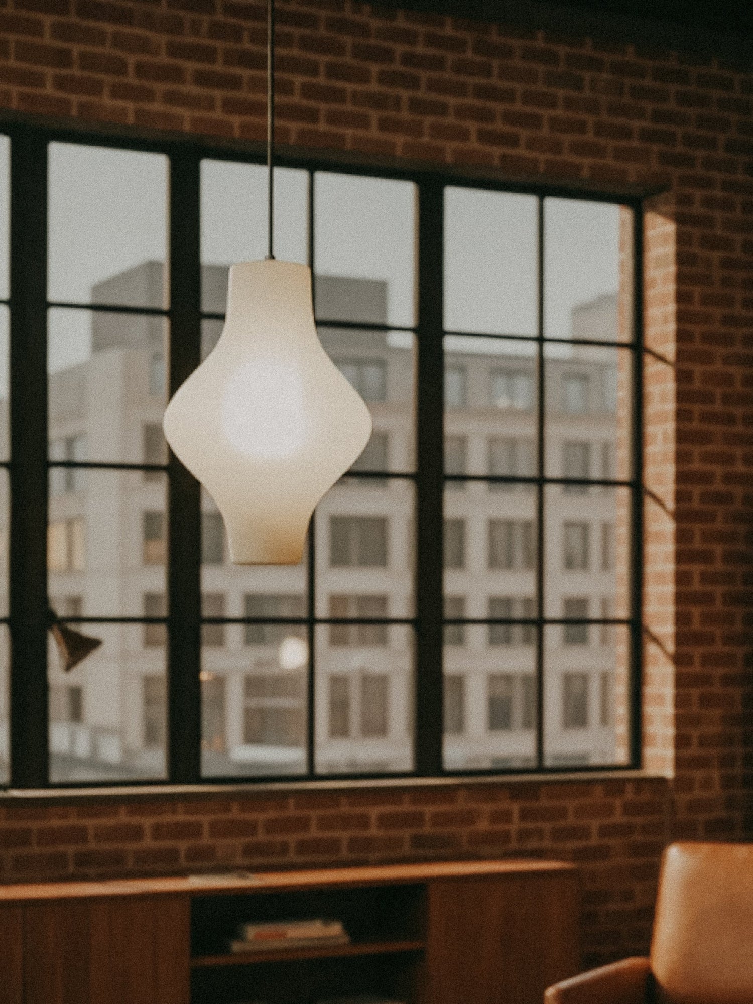 A modern white pendant lamp hangs in front of large industrial-style windows with a city building visible outside, inside a room with brick walls.