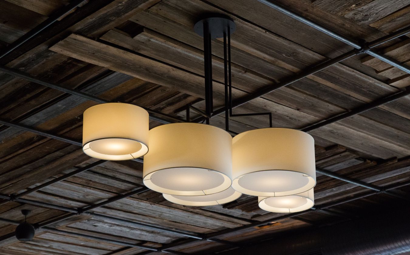 Modern chandelier with five cylindrical white lampshades hanging from a dark ceiling with exposed wooden beams.