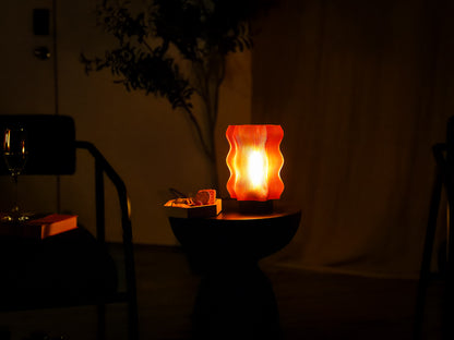 An OrijaDesign Reef Table Lamp, featuring wavy orange coral-inspired details, glows atop a small round table with a snack tray beside it; a glass of wine and a chair are set in the softly lit background, evoking a tranquil ocean decor atmosphere.