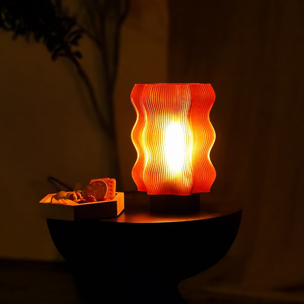 An OrijaDesign Reef Table Lamp, glowing with vibrant orange hues, rests on a round black table next to a small tray of assorted snacks, set against a dark, softly lit background.