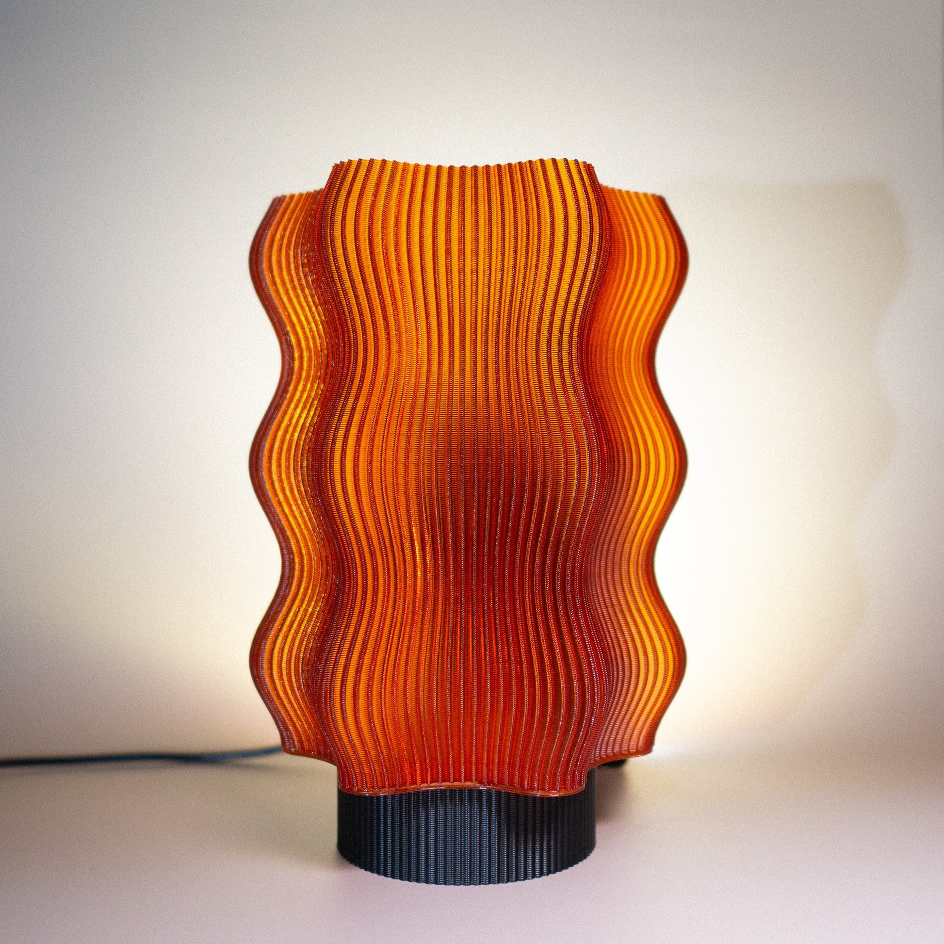 The OrijaDesign Reef Table Lamp features orange, wavy ribbed glass and a cylindrical black base, inspired by coral reefs for striking ocean-themed decor, shown illuminated against a white background.