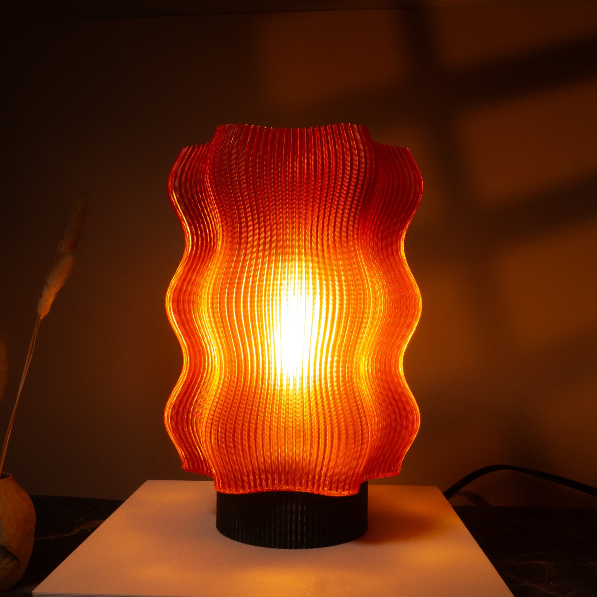 The Reef Table Lamp by OrijaDesign features a modern, wavy orange translucent shade that evokes coral reefs, casting warm light and soft shadows on surrounding surfaces.

