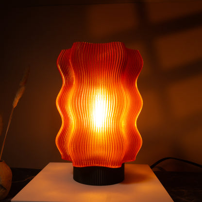 The Reef Table Lamp by OrijaDesign features a modern, wavy orange translucent shade that evokes coral reefs, casting warm light and soft shadows on surrounding surfaces.

