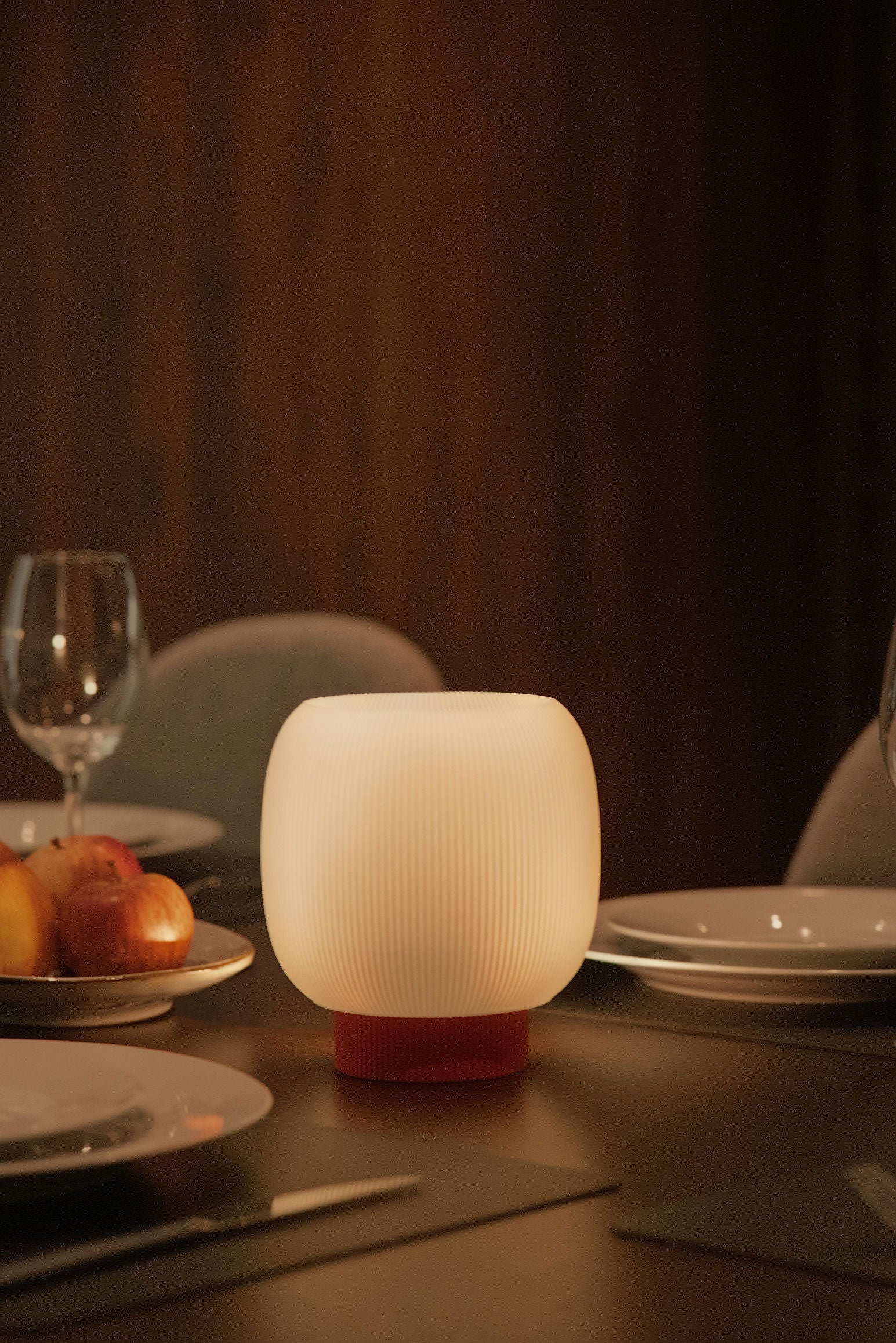 The Mochi Table Lamp by OrijaDesign, featuring a sculptural round, ribbed design, casts an ambient glow over a dining table set with plates, cutlery, a wine glass, and bread rolls.