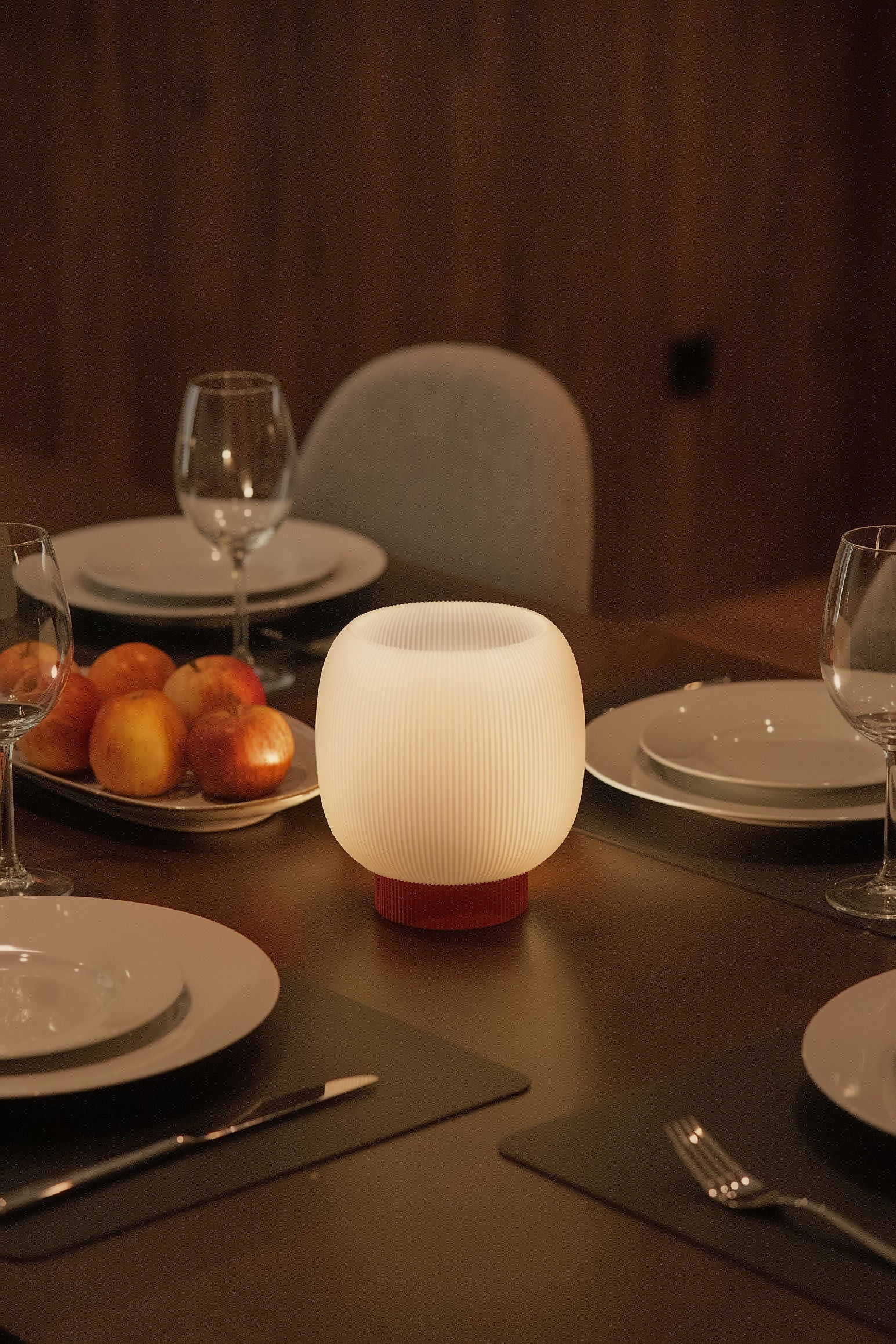 The Mochi Table Lamp by OrijaDesign sits on a dining table set with plates, glasses, cutlery, and a platter of apples in a warmly lit room, casting a gentle diffusion of light that enhances the ambient glow.