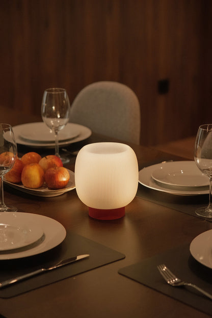 The Mochi Table Lamp by OrijaDesign sits on a dining table set with plates, glasses, cutlery, and a platter of apples in a warmly lit room, casting a gentle diffusion of light that enhances the ambient glow.