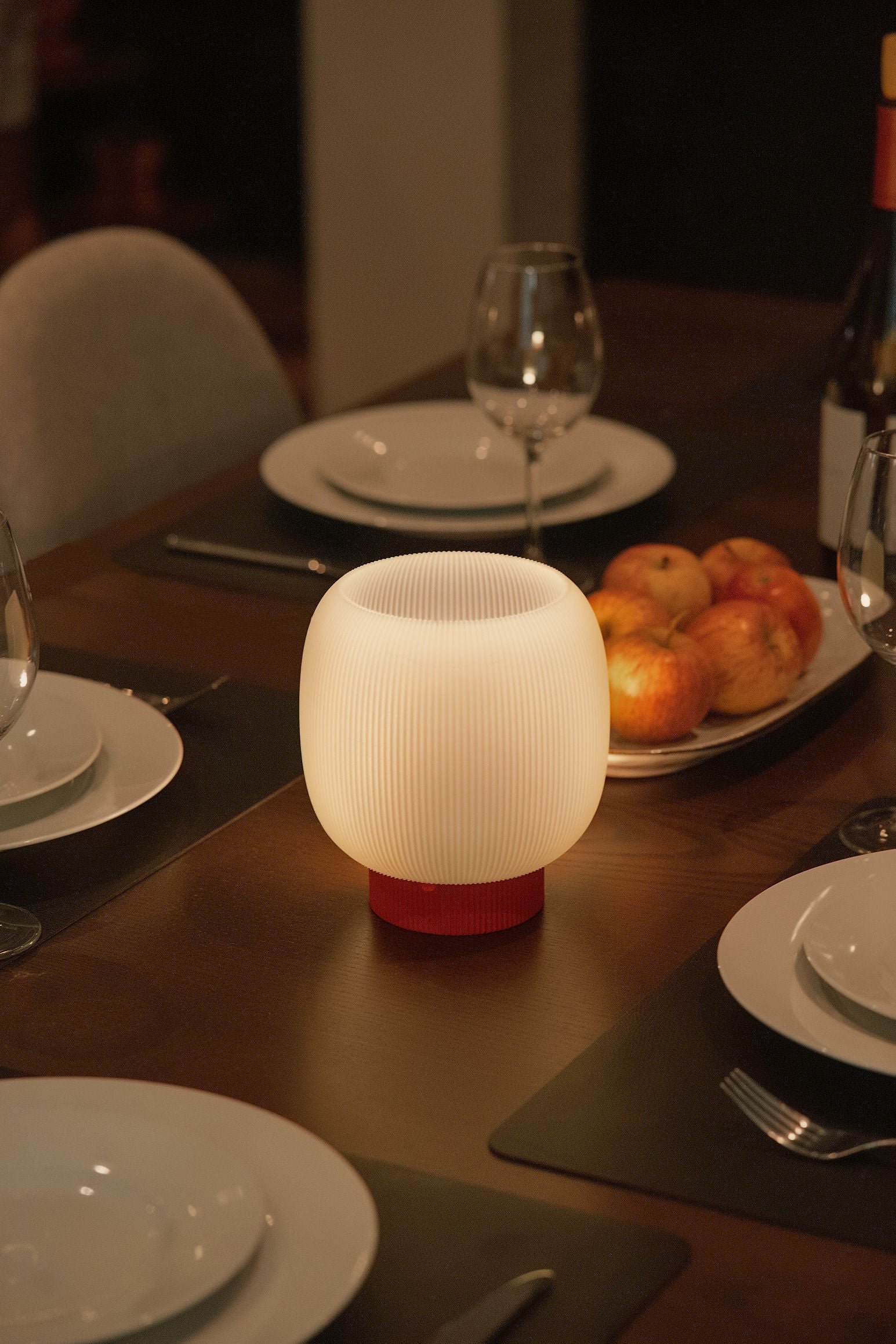 The Mochi Table Lamp by OrijaDesign, featuring a round, ribbed sculptural design and emitting a soft glow, sits on a dining table arranged with plates, glasses, and a fruit platter, casting an ambient light across the setting.