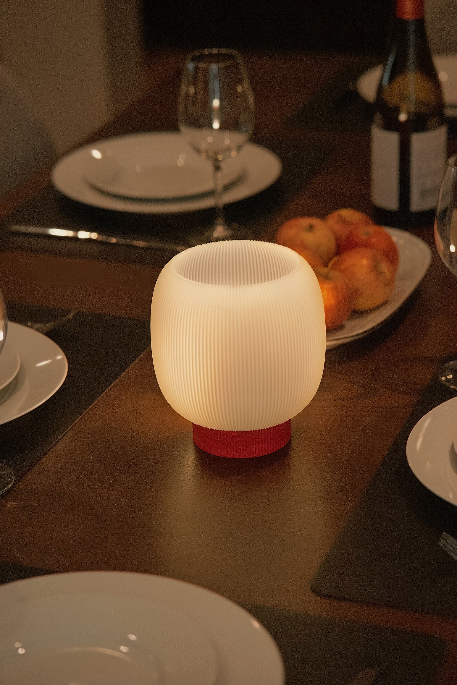 The Mochi Table Lamp by OrijaDesign, featuring a round, ribbed sculptural base and a glowing white shade, stands on a set dinner table arranged with plates, glasses, and a bowl of apples. It casts an ambient glow and gently diffuses light across the elegant setting.