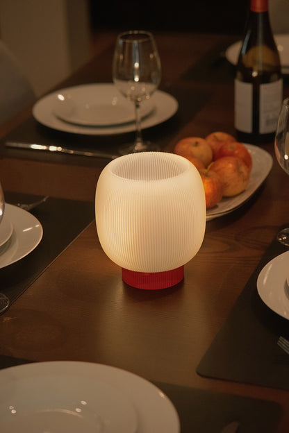 The Mochi Table Lamp by OrijaDesign, featuring a round, ribbed sculptural base and a glowing white shade, stands on a set dinner table arranged with plates, glasses, and a bowl of apples. It casts an ambient glow and gently diffuses light across the elegant setting.