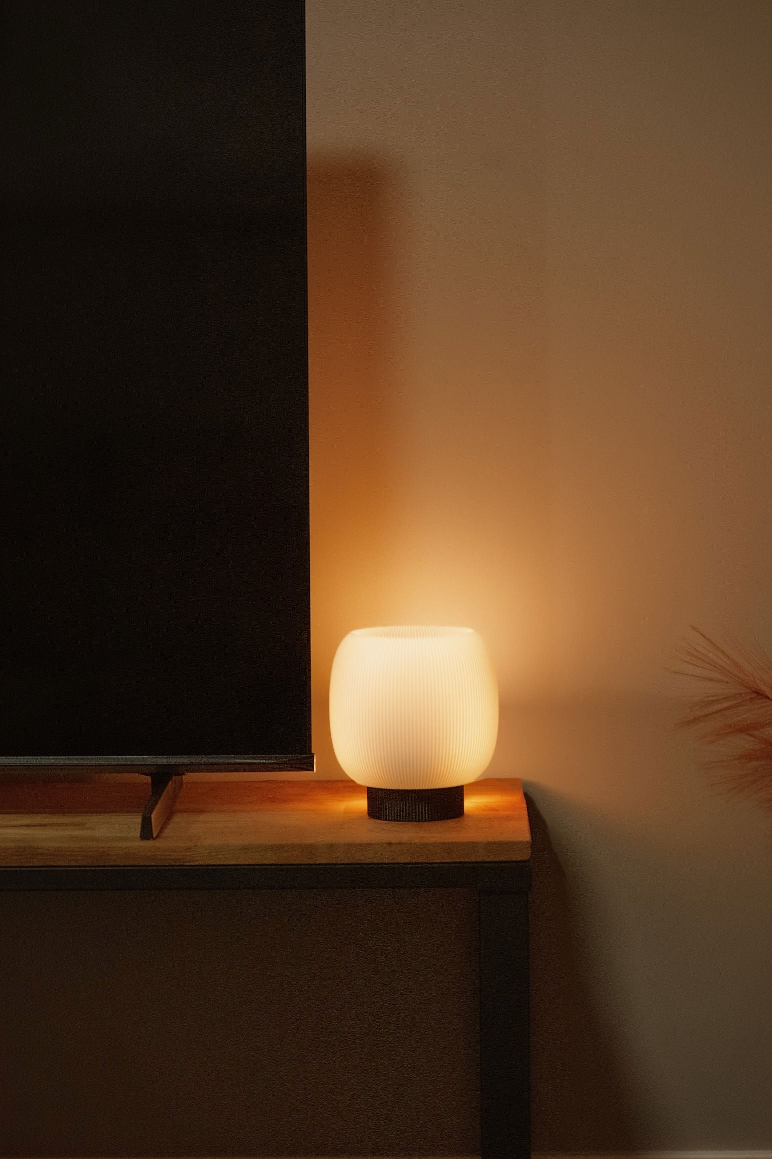 The Mochi Table Lamp by OrijaDesign sits on a wooden table next to a flat-screen TV against a beige wall, its round, sculptural form gently diffusing light and casting an ambient glow.