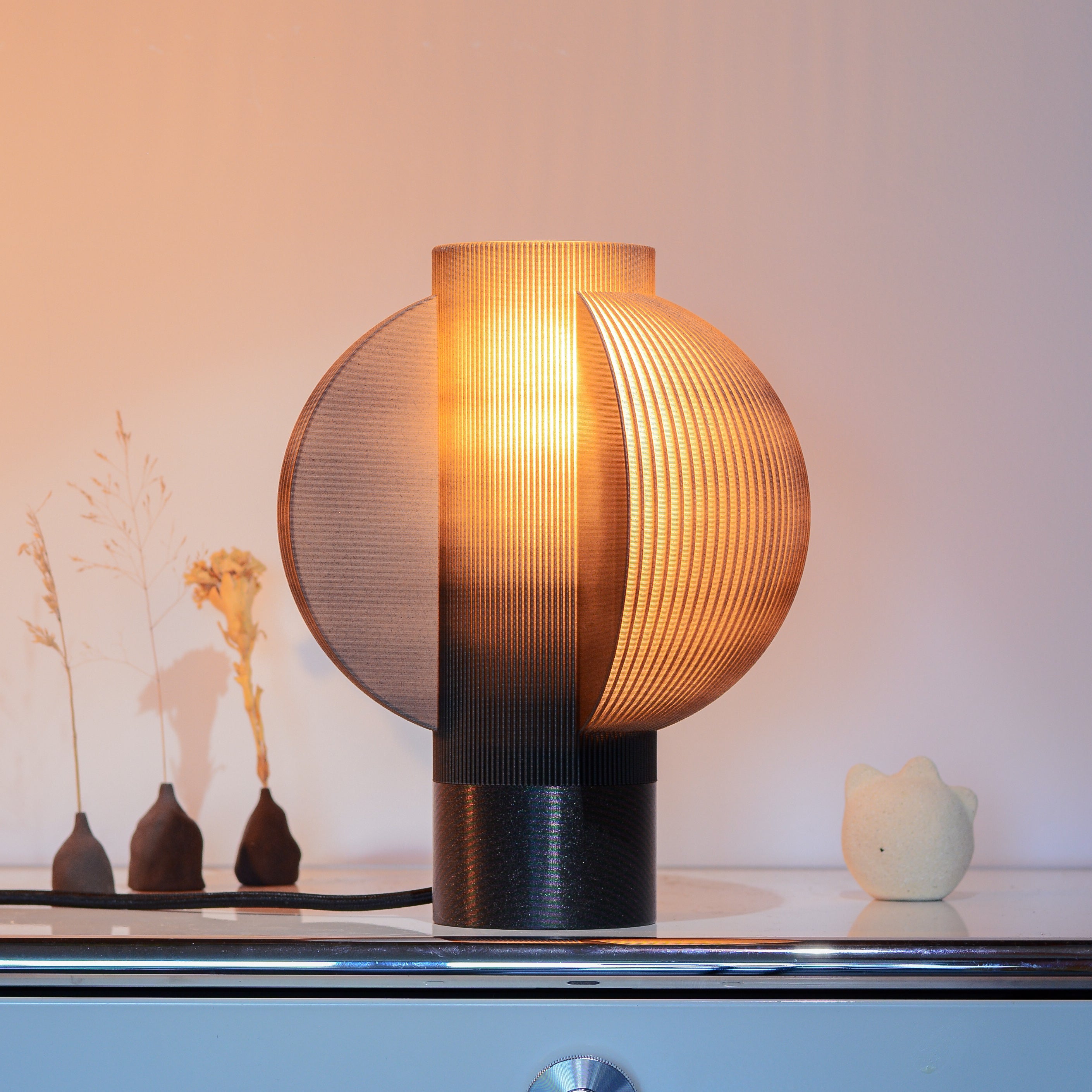 The Lemon Table Lamp - Small emits a warm glow on a sleek surface. Dried flowers in vases and a decorative object in the background enhance the ambiance, making it an ideal bedside light with its modern black design.