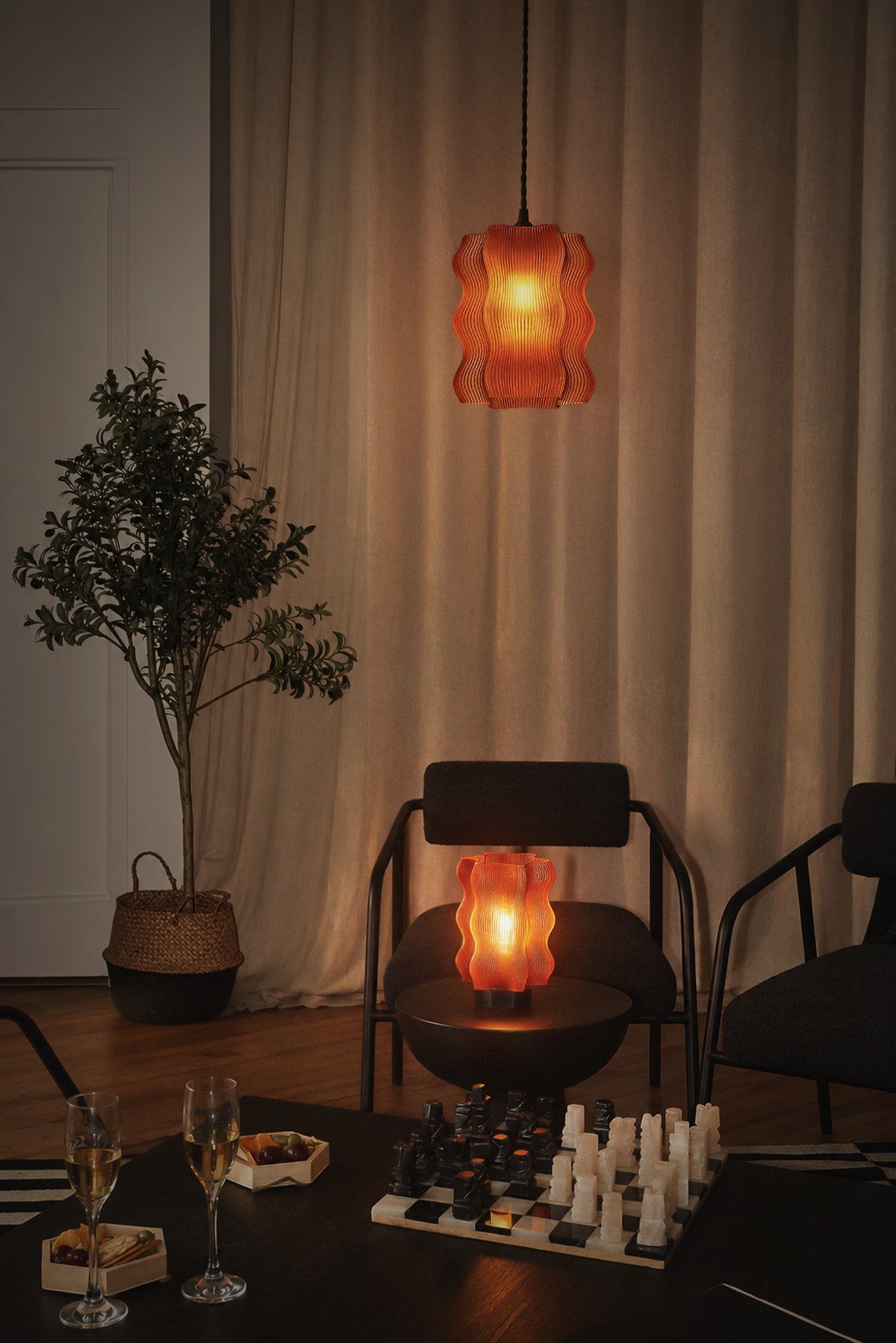 A cozy living room features two Reef Pendant lamps by My Store that cast an atmospheric glow, a potted plant, two black chairs, a chess set, two glasses of wine, and snacks on a dark table, with beige curtains in the background.