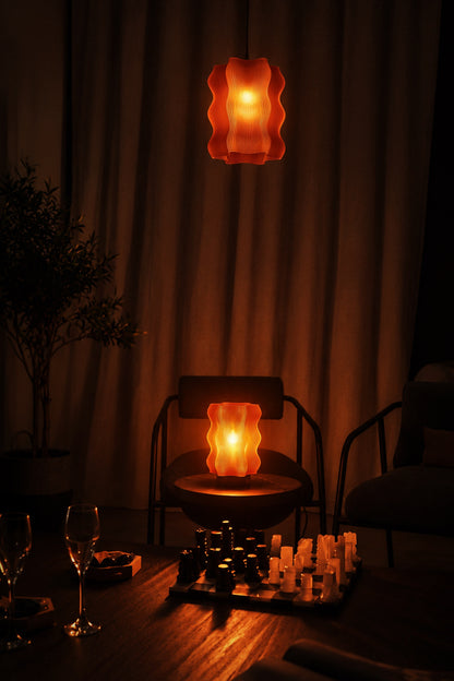 A dimly lit room featuring two orange wavy lamps casting an atmospheric glow, the Reef Pendant by My Store hanging above a chair, a potted plant, a chessboard, and wine glasses on the table.