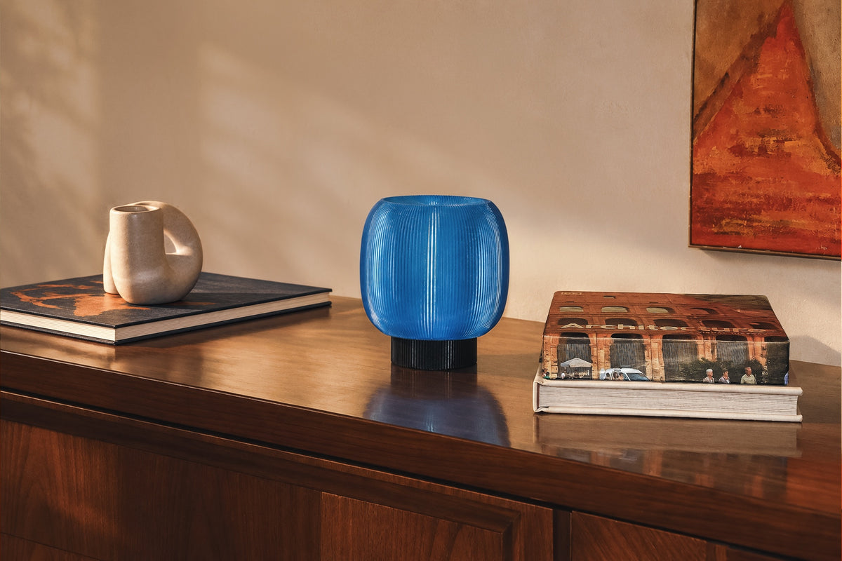 A rounded, blue, ribbed Mochi Table Lamp by OrijaDesign with a dark base sits on a wooden cabinet, providing a gentle diffusion of light between two stacks of books and a sculpture.