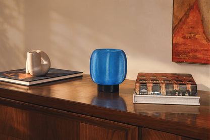 A rounded, blue, ribbed Mochi Table Lamp by OrijaDesign with a dark base sits on a wooden cabinet, providing a gentle diffusion of light between two stacks of books and a sculpture.