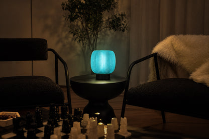 A glowing turquoise, ribbed glass Mochi Table Lamp by OrijaDesign provides an ambient glow as it sits on a black pedestal table between two chairs in a dimly lit room.