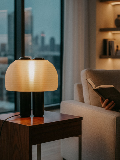 A person reads on a sofa near a wooden side table topped with the OrijaDesign Nami Table Lamp, featuring a pleated lampshade, as a cityscape is visible through the window in the background.