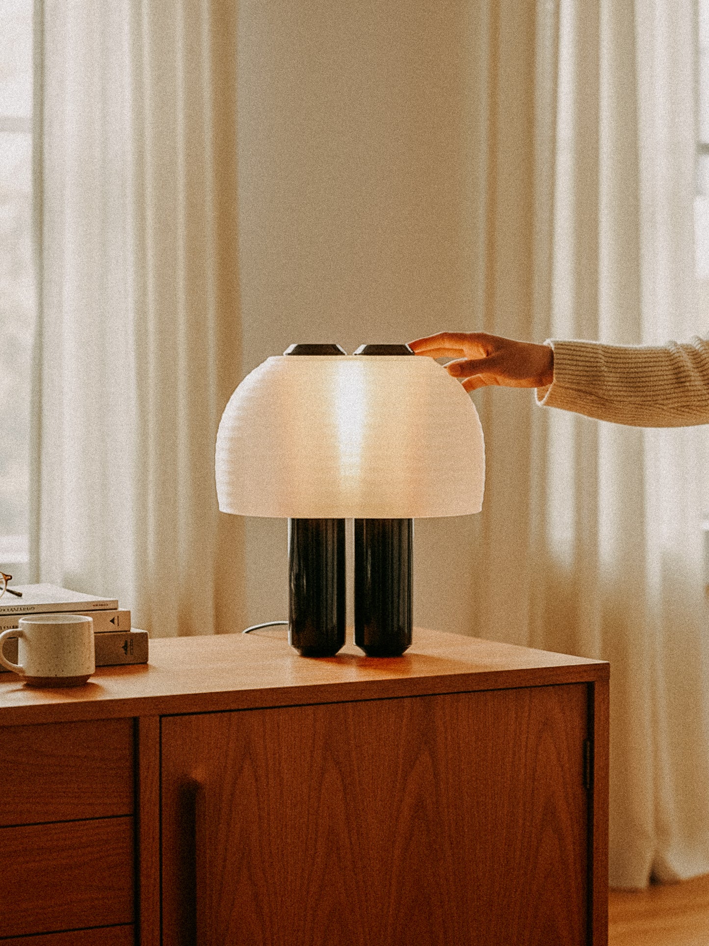A person reaches to touch the OrijaDesign Nami Table Lamp, featuring a pleated lampshade inspired by Japanese paper lanterns, placed on a wooden cabinet in a softly lit room with curtains and a cup.