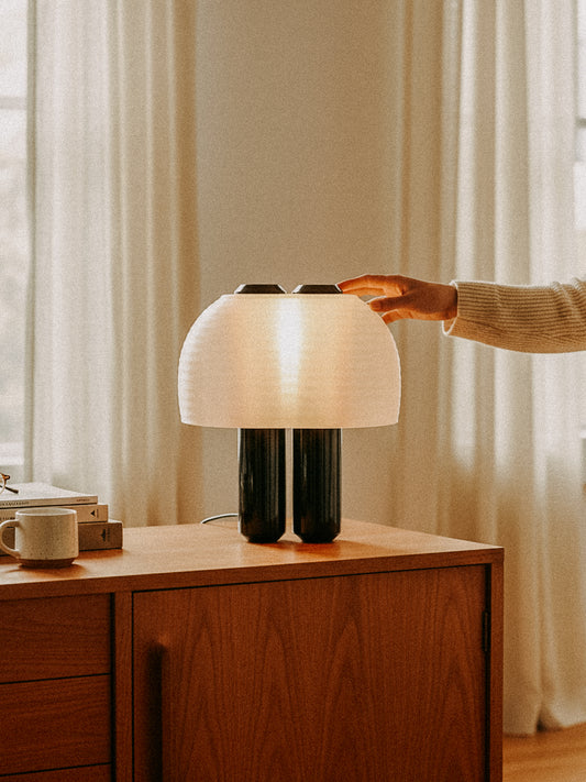 A person reaches to touch the OrijaDesign Nami Table Lamp, featuring a pleated lampshade inspired by Japanese paper lanterns, placed on a wooden cabinet in a softly lit room with curtains and a cup.