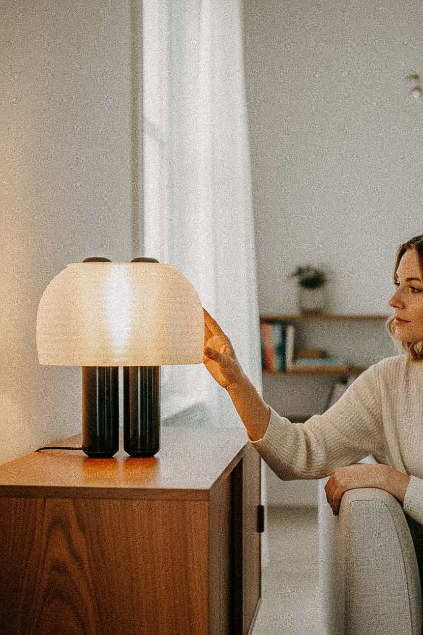A woman on a couch reaches for the OrijaDesign Nami Table Lamp, featuring a pleated lampshade, placed on a wooden sideboard in a softly lit room.