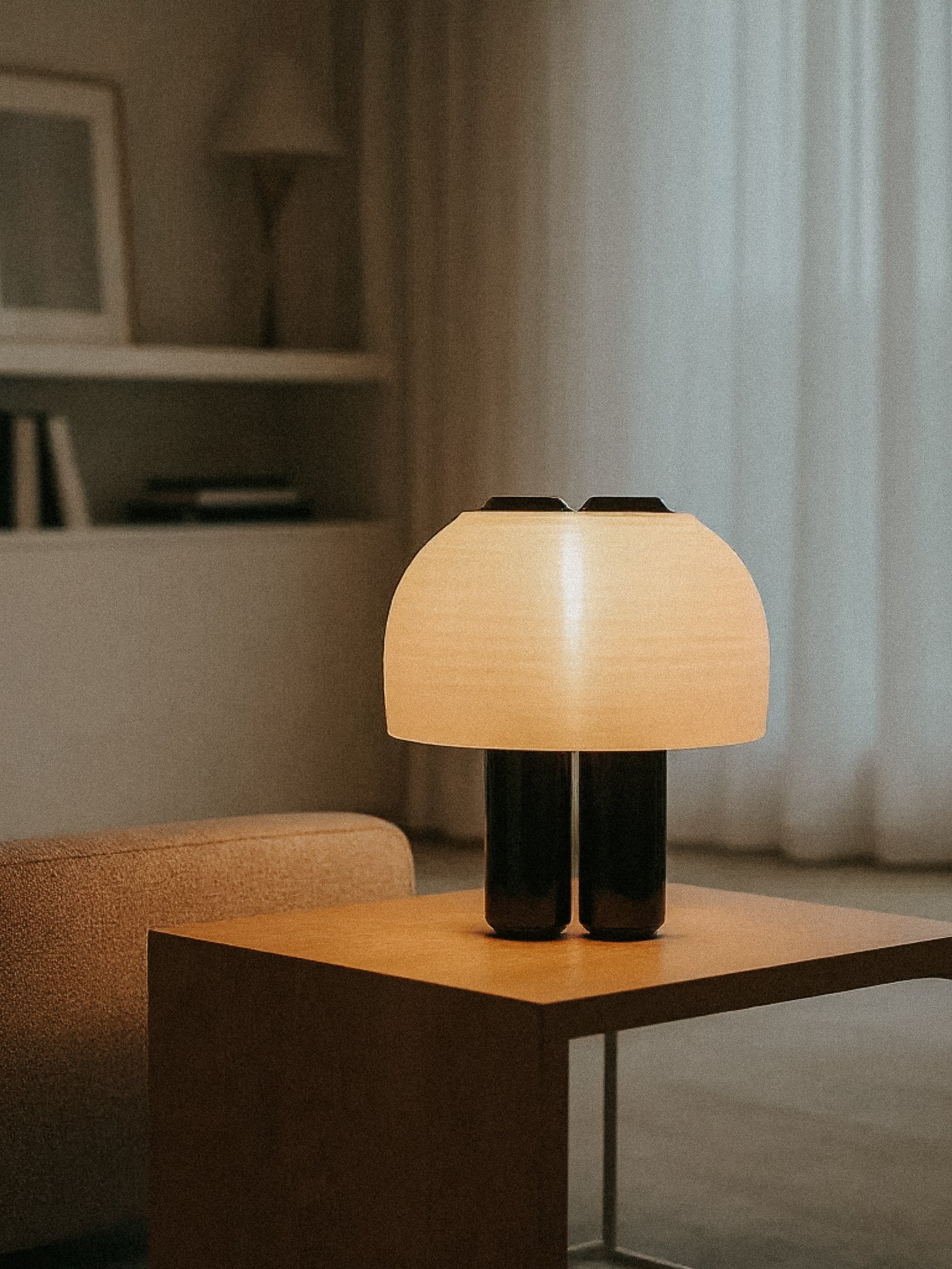 The OrijaDesign Nami Table Lamp, featuring a pleated lampshade, rests on a wooden side table in a softly lit minimalist living room, capturing the elegance of Japanese paper lanterns.