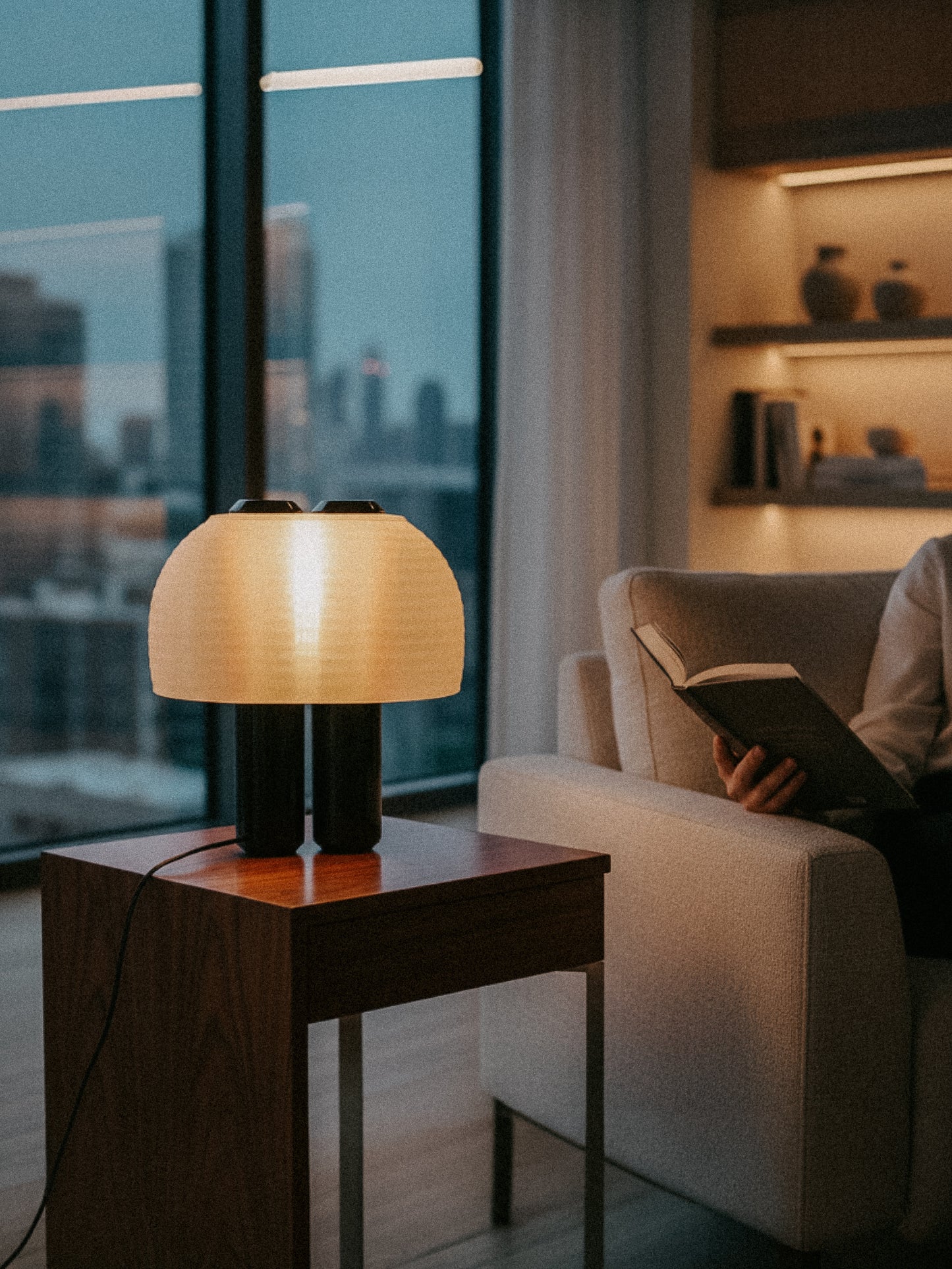 An OrijaDesign Nami Table Lamp with a pleated lampshade rests on a wooden side table by a beige armchair, where someone is reading in a softly lit living room with large windows.