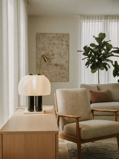 A modern living room with neutral furniture, a large plant, sheer curtains, and abstract art, featuring the OrijaDesign Nami Table Lamp with a pleated shade inspired by Japanese paper lanterns.