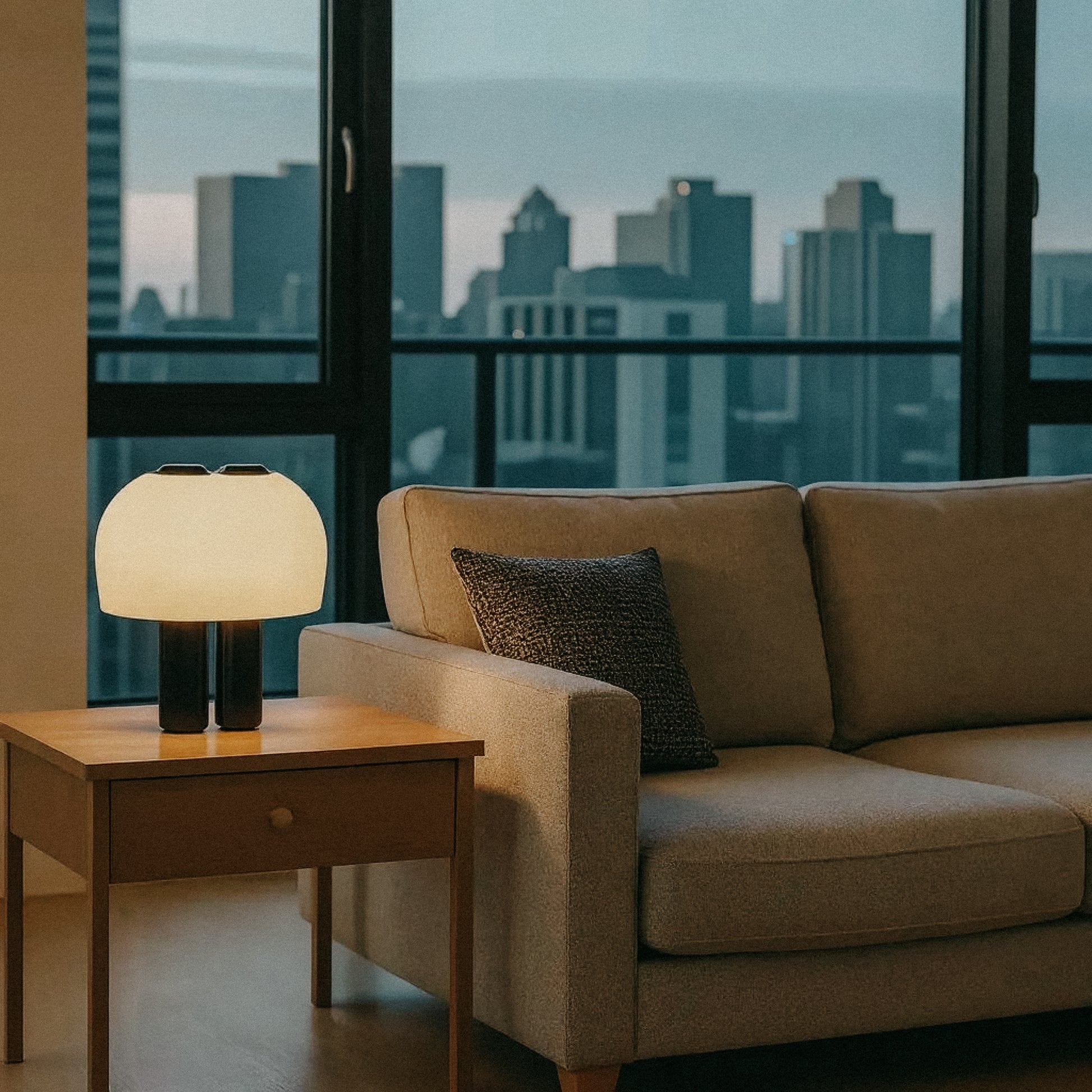 A modern living room with a beige sofa and side table topped with OrijaDesign's Nami Table Lamp, featuring a pleated lampshade, set before large windows overlooking the city skyline at dusk.