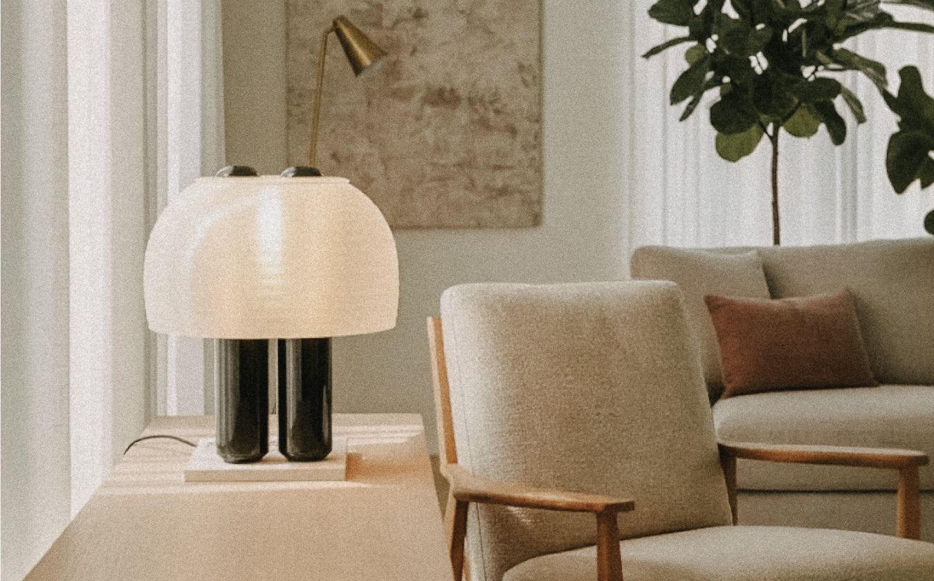 A modern table lamp with a white shade sits on a wooden side table next to a gray armchair, with a sofa, throw pillow, plant, and wall art in the background.