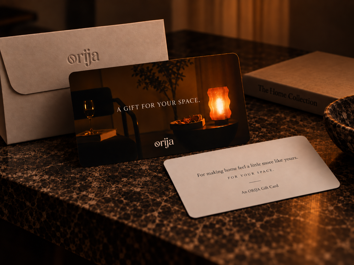 A Gift Card and envelope from OrijaDesign Store rest on a marble table beside a bowl, illuminated by warm ambient lighting. The card reads: “A GIFT FOR YOUR SPACE.”.