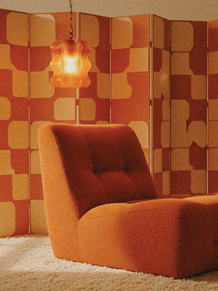 A retro lounge chair and the My Store Reef Pendant with brass hardware hang in front of an orange and yellow geometric room divider on a white shag carpet.