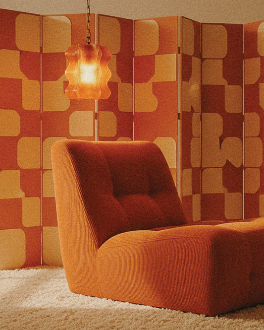 A retro lounge chair and the My Store Reef Pendant with brass hardware hang in front of an orange and yellow geometric room divider on a white shag carpet.