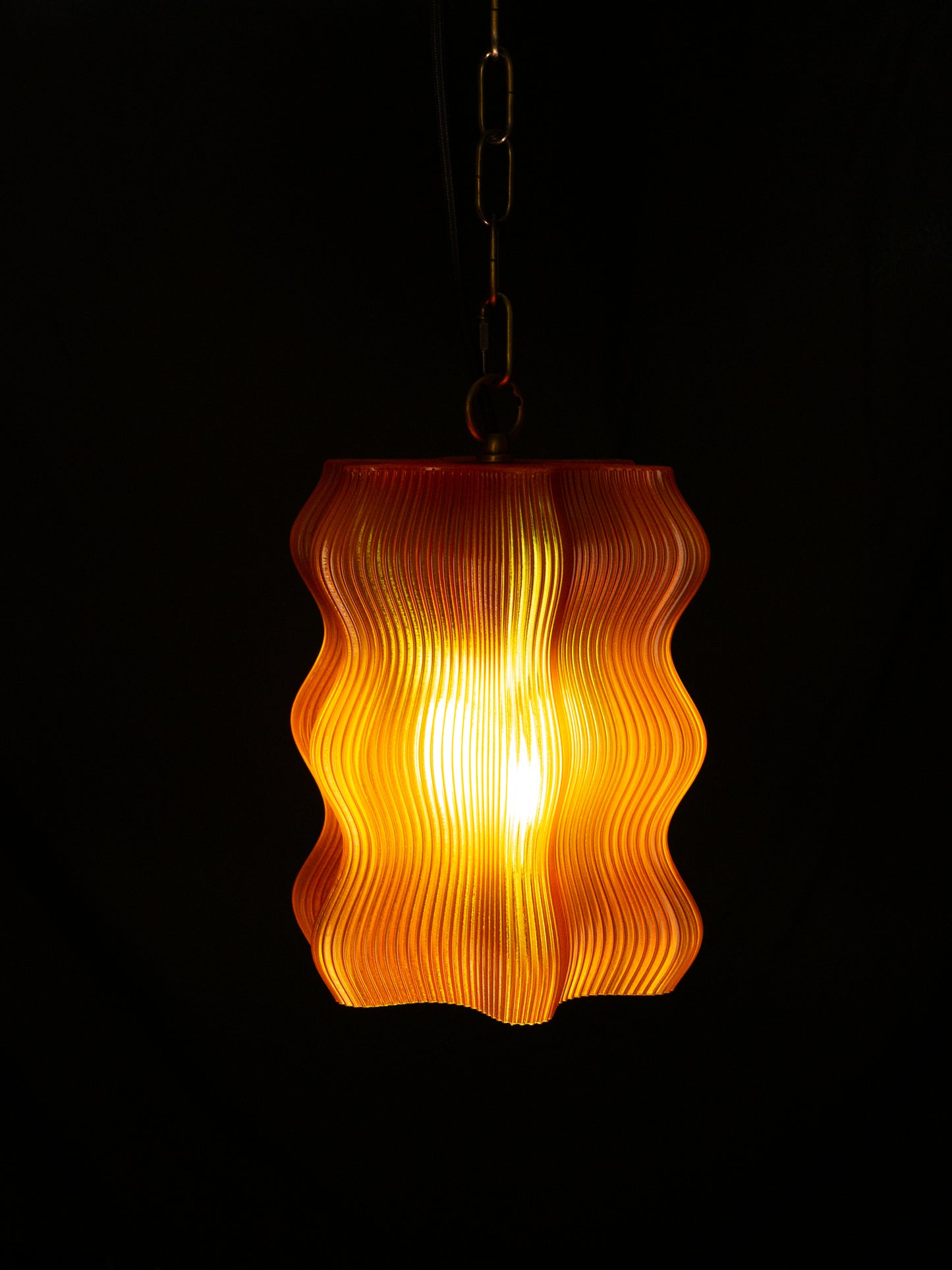 The My Store Reef Pendant features a wavy orange shade suspended from brass hardware, shown illuminated against a black background.