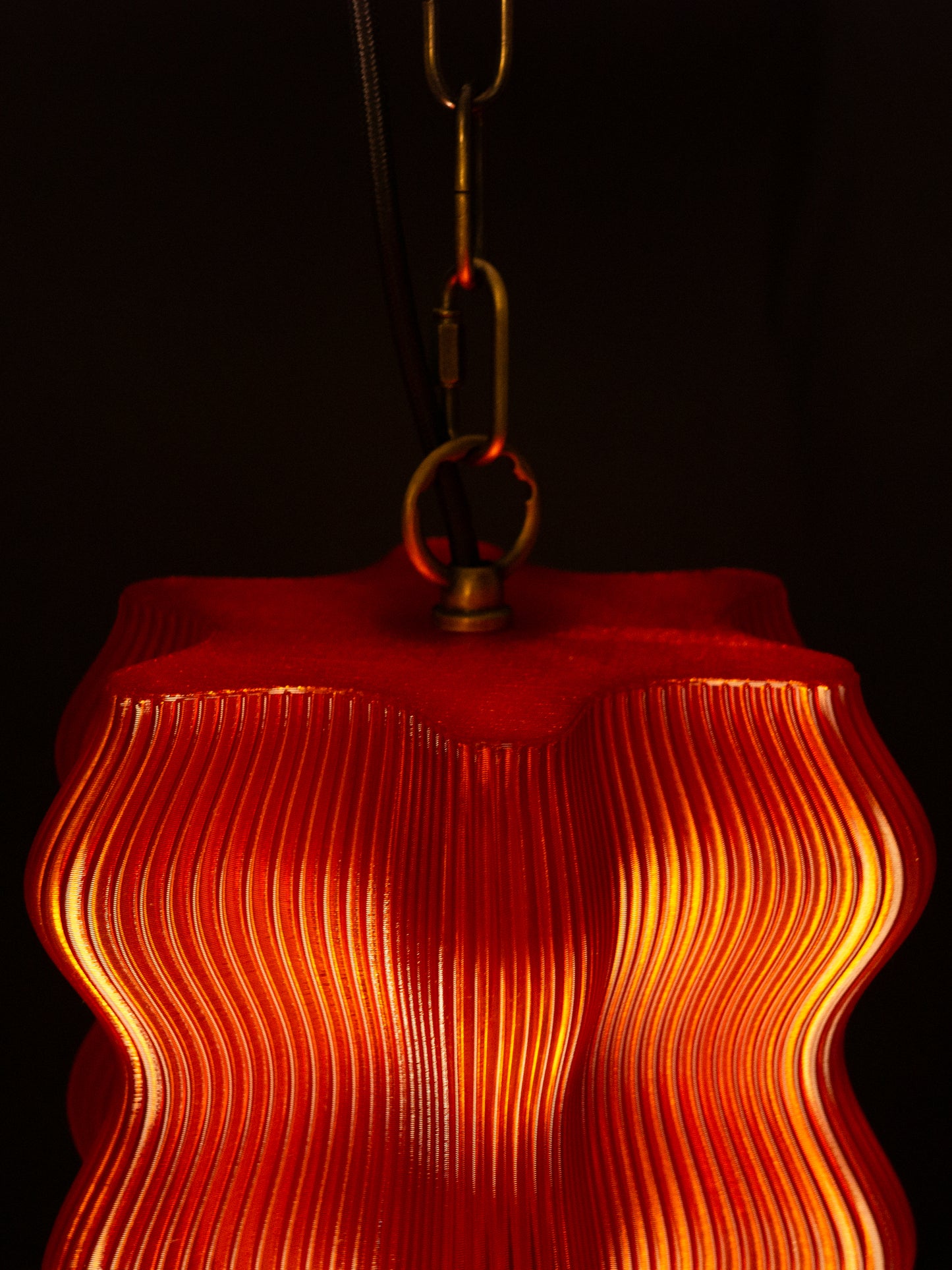 The Reef Pendant by My Store features a wavy, ribbed red shade and brass hardware, casting a warm yellow glow against a dark backdrop.