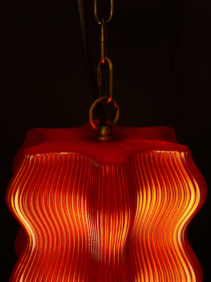 The Reef Pendant by My Store features a wavy, ribbed red shade and brass hardware, casting a warm yellow glow against a dark backdrop.