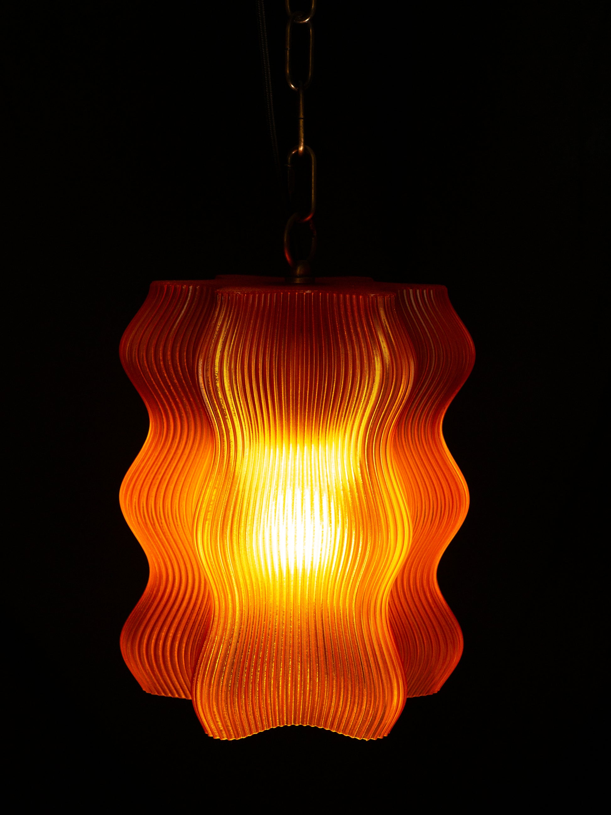 The Reef Pendant by My Store features a glowing orange wavy, ribbed shade with elegant brass hardware, casting a warm light against a dark background.