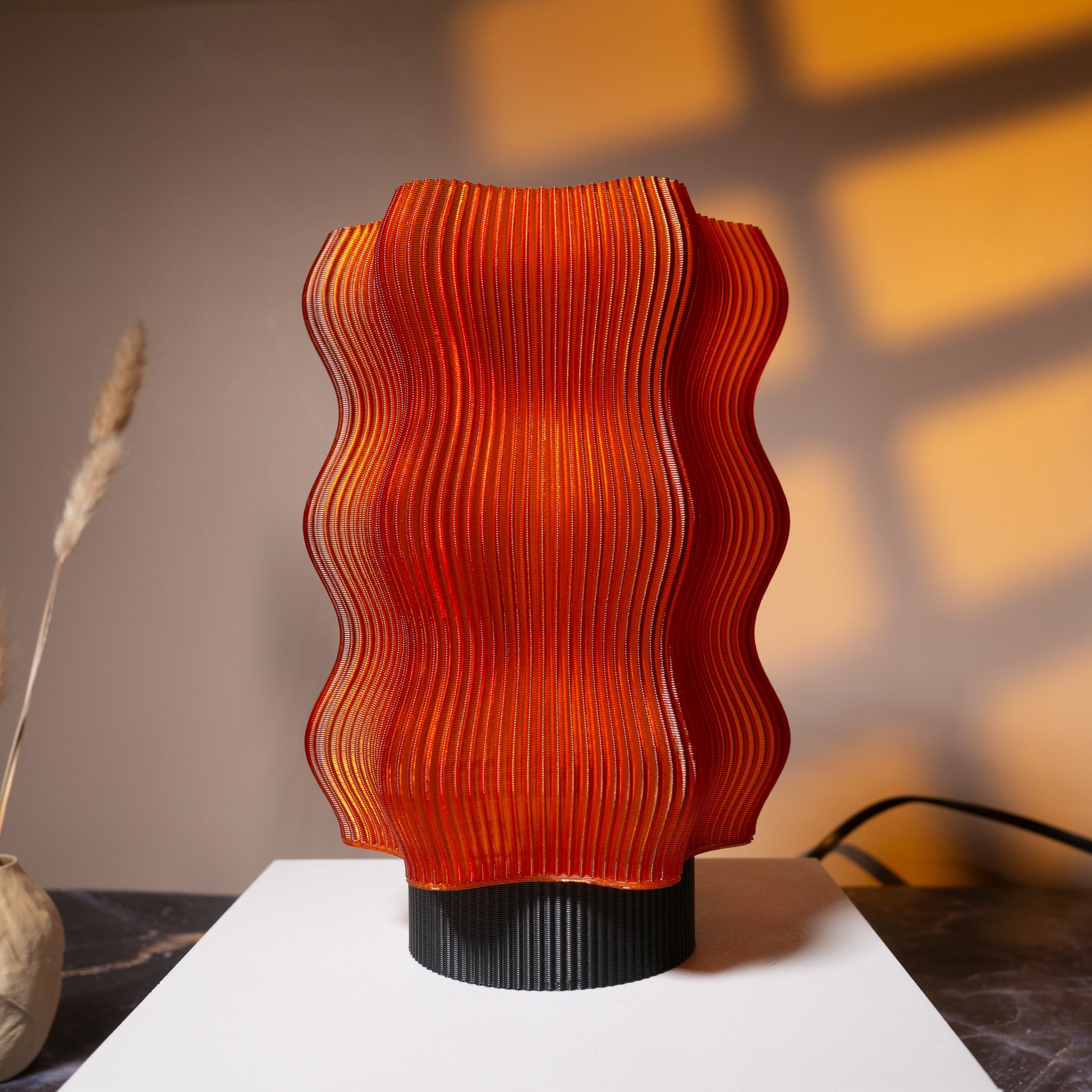 The OrijaDesign Reef Table Lamp (Seconds) features a wavy, ribbed red-orange shade inspired by coral reefs. It stands on a white surface, casting warm light and shadows—a vibrant accent for your decor.