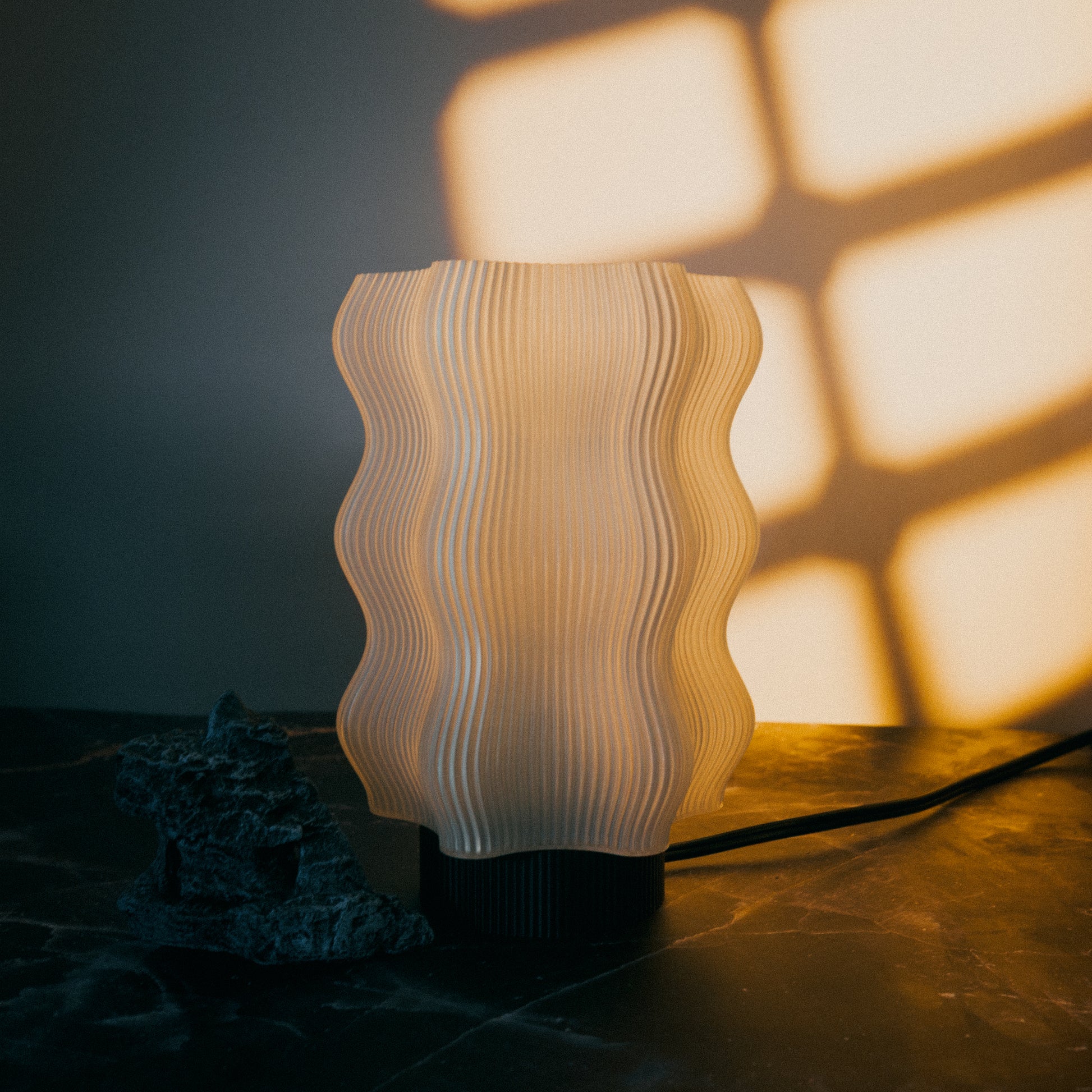 The Reef Table Lamp by OrijaDesign glows warmly on a dark surface near a rough stone, evoking coral reefs’ textured beauty and casting geometric shadows on the wall.