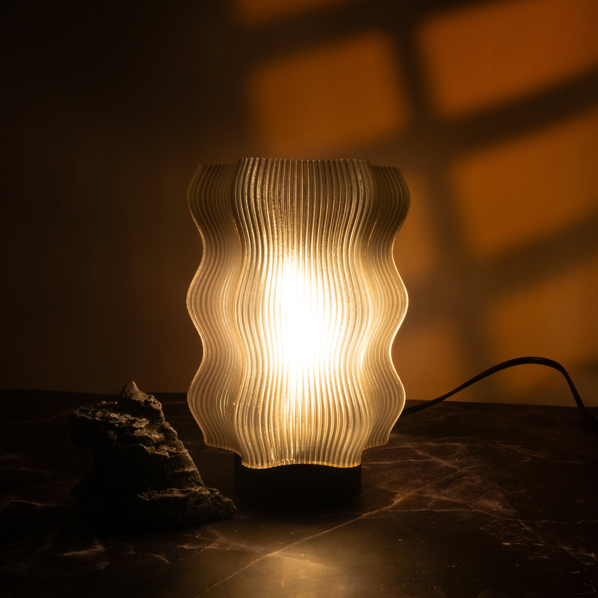 The Reef Table Lamp (Seconds) by OrijaDesign emits warm, wavy light that casts coral reef-like shadows on the wall. A nearby dark rock on the surface enhances the tranquil decor.