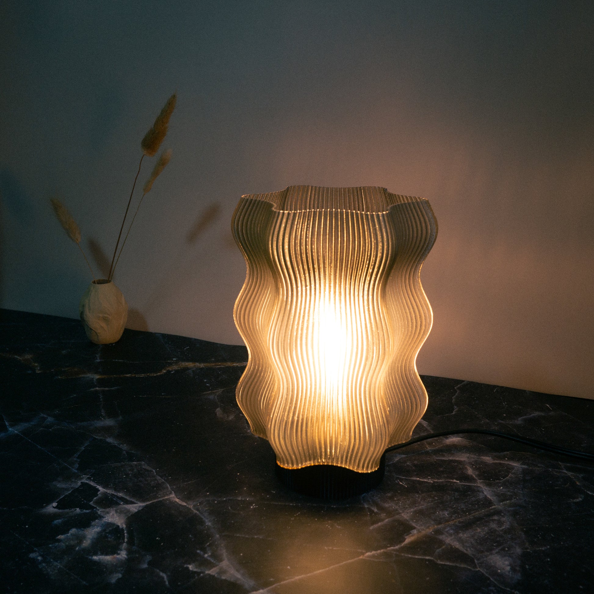 The Reef Table Lamp by OrijaDesign, with its wavy ribbed design, casts warm light over a dark marble surface, evoking ocean decor. A small vase with dried grasses sits in the background.