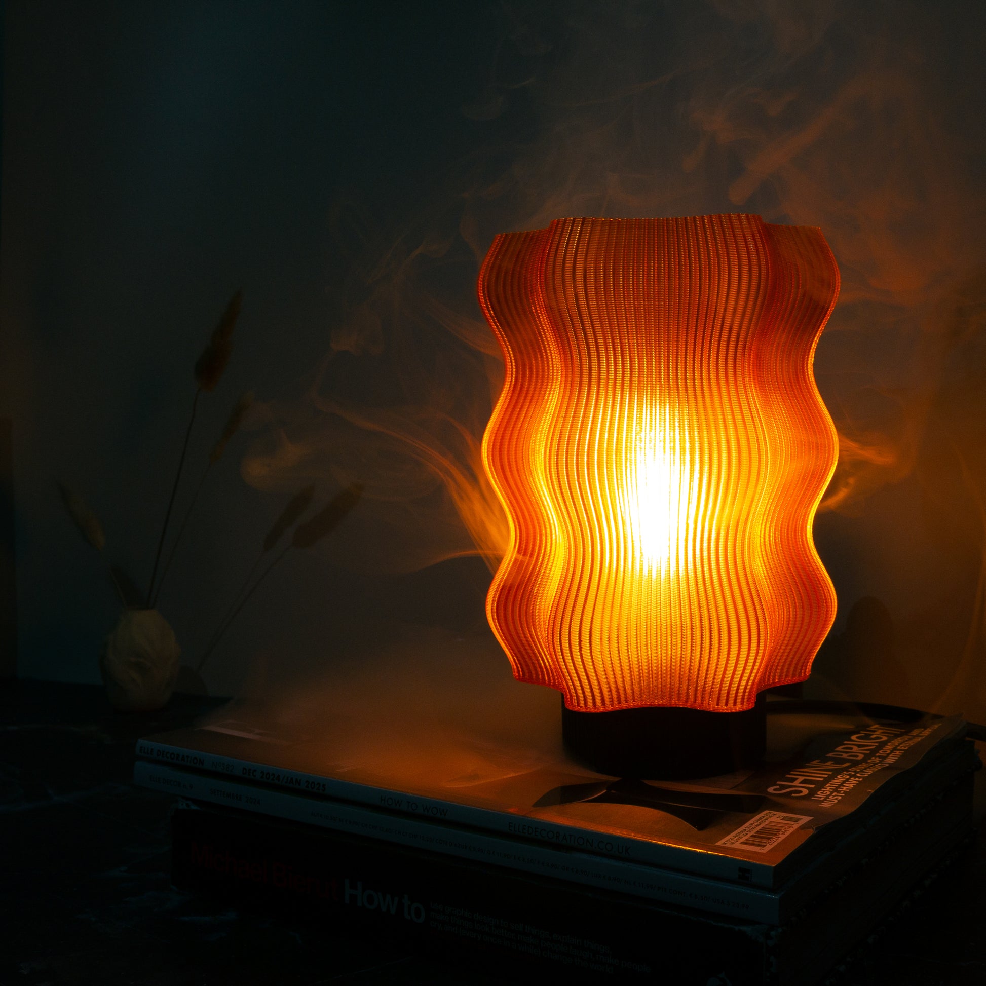 The Reef Table Lamp (Seconds) by OrijaDesign features a wavy, ribbed orange design and emits a warm glow—an eye-catching decor piece inspired by vibrant coral reefs, perfect for adding charm to any room.