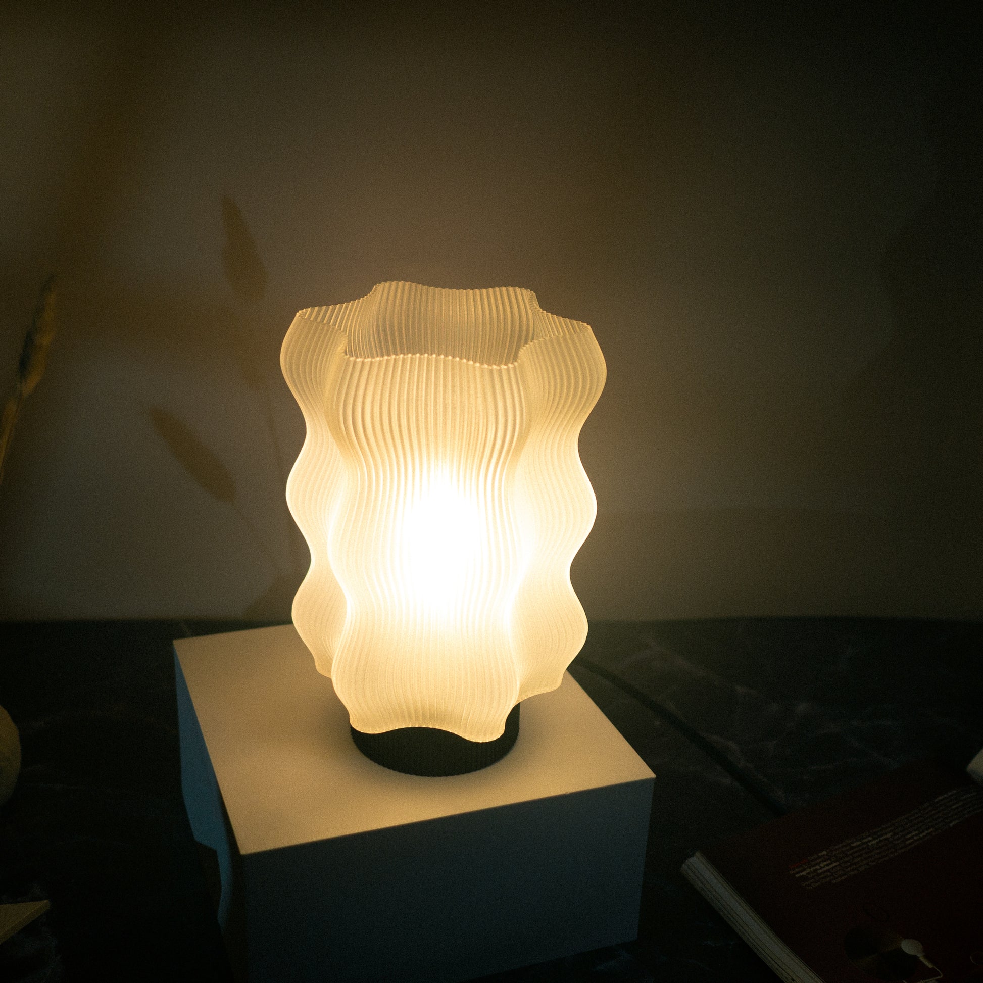The Reef Table Lamp by OrijaDesign features a wavy white base and ribbed shade, bringing ocean-inspired style and a soft glow to dark surfaces. A book rests partially visible nearby.