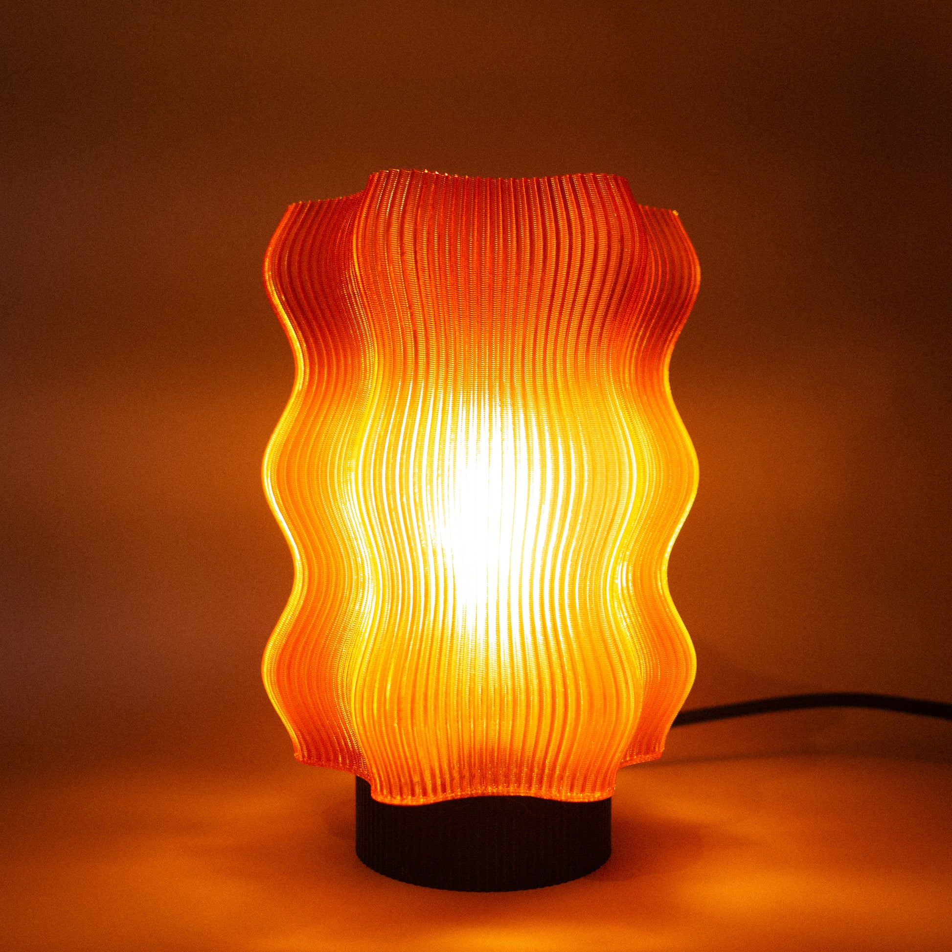 The OrijaDesign Reef Table Lamp (Seconds) features a wavy orange ribbed shade inspired by coral reefs, casting a warm yellow glow and adding distinctive decor to any space.