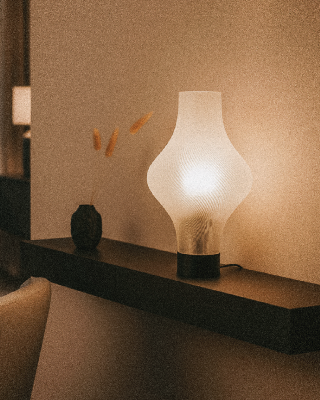 The OrijaDesign Solaris Table Lamp, featuring a modern sculptural white shade, rests on a dark floating shelf beside a vase with dried grass, offering elegant bedside lighting in a softly lit room.