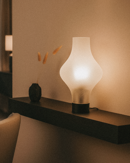 The OrijaDesign Solaris Table Lamp, featuring a modern sculptural white shade, rests on a dark floating shelf beside a vase with dried grass, offering elegant bedside lighting in a softly lit room.